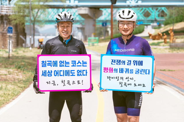 김태경 칠곡군 사이클연맹 회장(왼쪽)과 김원일 원바이오젠 대표가 칠곡 자전거길에서 캠페인 피켓을 들고 기념촬영을 하고 있다.  두 사람은 ‘칠곡에 없는 코스는 세상 어디에도 없다’,‘전쟁의 길 위에 평화의 바퀴를 굴린다’는 문구로 자전거 도시 칠곡의 상징성과 그란폰도 대회의 의미를 전했다. /칠곡군제공